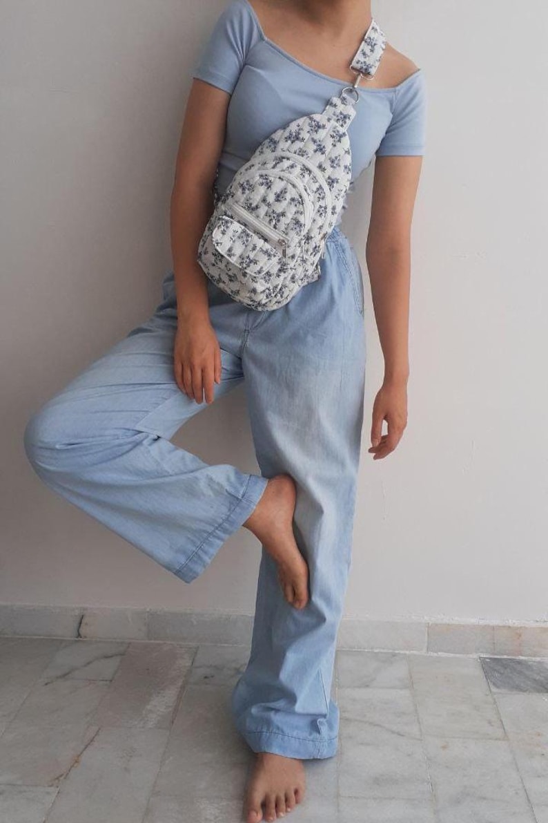 May include: A white sling bag with a blue floral pattern is worn over the shoulder. The bag has a small front pocket and a matching strap. The person is wearing a light blue top and light blue pants.