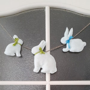 Op de afbeelding: Een slinger van drie witte vilt konijnen met groene en blauwe strikken die aan een witte deur met een glazen paneel hangen.