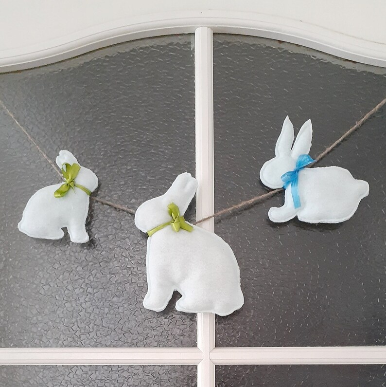 Op de afbeelding: Een slinger van drie witte vilt konijnen met groene en blauwe strikken die aan een witte deur met een glazen paneel hangen.
