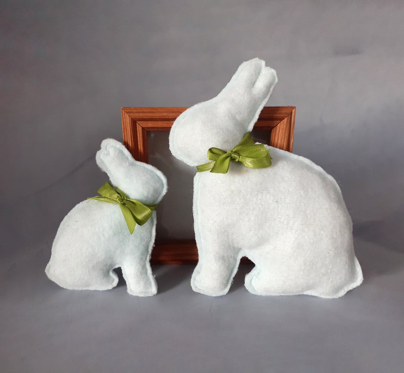 Op de afbeelding: Twee pluche lichtblauwe konijnen met groene strikken. Het grotere konijn zit voor een houten fotolijst.