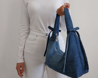 Op de afbeelding: Een denim tas met strik, een ritssluiting en een lange schouderband. De tas wordt vastgehouden door iemand die een witte broek en een wit lang mouw shirt draagt. BOW BAG PATTERN staat in zwarte tekst onderaan de afbeelding.