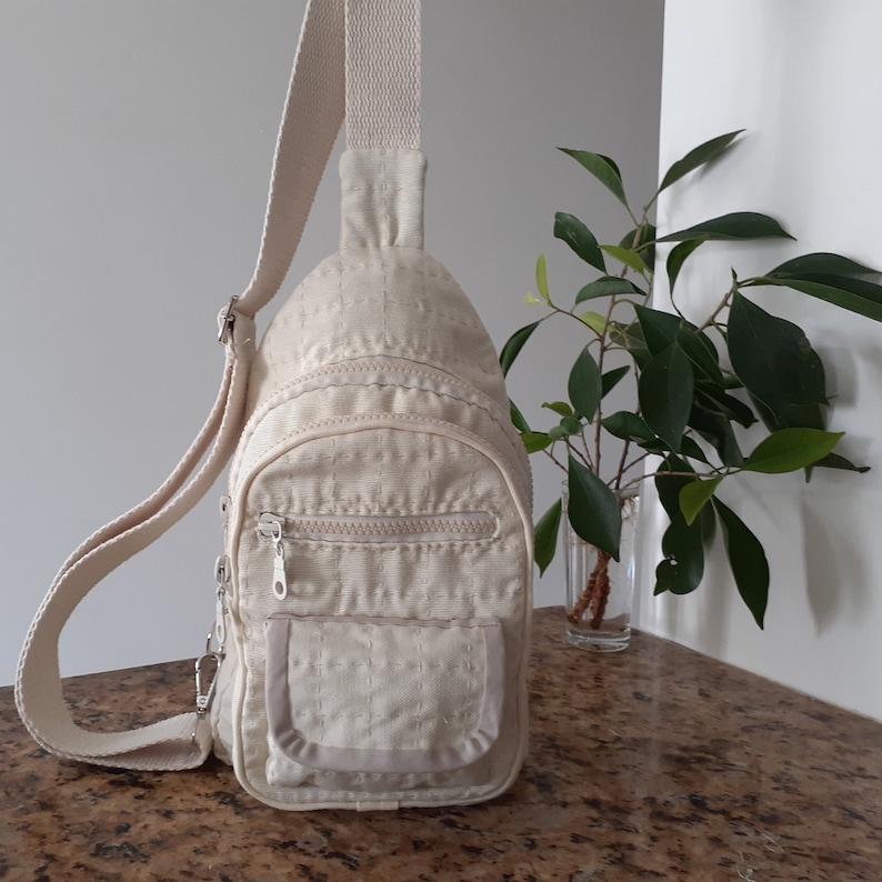 May include: Beige sling bag with a quilted pattern, featuring a zippered main compartment and a smaller front pocket. The bag has a long, adjustable strap and silver-tone hardware. The bag is made of a durable, lightweight material.