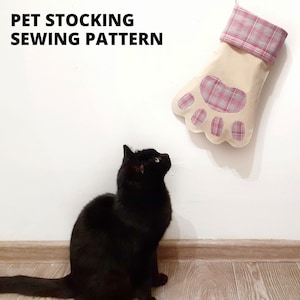 Puede incluir: Un gato negro mira hacia arriba a una media beige en forma de pata con un puño de cuadros rojos y blancos. La media está colgada en una pared blanca. El texto "PET STOCKING SEWING PATTERN" está en la parte superior de la imagen.