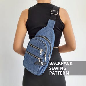 Op de afbeelding: Een blauwe denim sling bag met meerdere vakken met ritssluiting en een zwarte band. Een kleine zwarte vleermuisvormige bedel hangt aan de rits. De tekst "BACKPACK SEWING PATTERN" is zichtbaar in een witte doos.