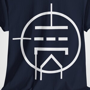 Puede incluir: Camiseta azul marino con un diseño gráfico blanco. El diseño es un símbolo circular estilizado con líneas horizontales y verticales, y una forma triangular en la parte inferior. La camiseta está hecha de un material suave.