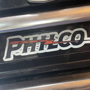May include: Black sticker with the word "PHILCO" in white, outlined in black, with a red lightning bolt design. The sticker is on a black surface with a silver metal handle.