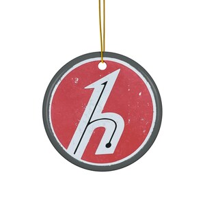 Puede incluir: Un adorno redondo de cerámica con un diseño rojo y blanco. El adorno presenta una letra "h" estilizada con un contorno negro y un interior blanco. El adorno tiene un aspecto desgastado, lo que le da un toque vintage.