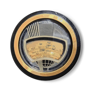 Peut inclure: Cadran de radio vintage, de forme circulaire, avec un bord noir et doré. Le cadran crème affiche des noms de stations comme Londres et Berlin, et le texte "MUSIC NEWS DRAMA COMOR". Il indique les fréquences.