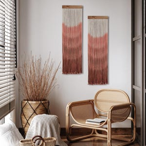 May include: Two rectangular wall hangings with a gradient design in shades of white, pink, and brown. The hangings are made of vertical strands attached to a wooden bar. A woven chair and a basket with tall dried grass are also visible.