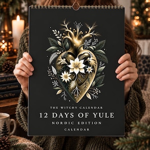 May include: A black wall calendar titled The Witchy Calendar 12 Days of Yule Nordic Edition. The cover features a central gold heart-shaped illustration adorned with white poinsettia flowers, pine branches, and delicate golden twigs.