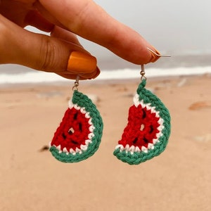 May include: Crochet earrings in the shape of watermelon slices. The earrings have a red centre with black seed details, a white rind, and a green outer edge. The earrings are suspended from silver hooks.