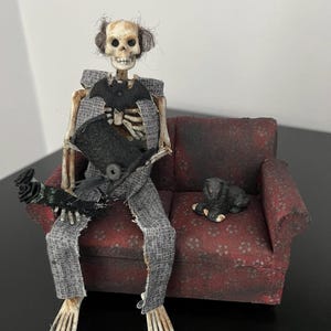 May include: A skeleton wearing a gray suit and a black top hat sits on a red couch with a black dog figurine.