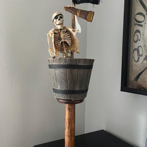 May include: A decorative skeleton figure wearing an eye patch and tattered clothing, positioned inside a wooden barrel with black bands. The skeleton holds a brown telescope and stands next to a small black flag featuring a white skull and crossbones.