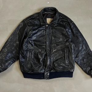 Può includere: Giacca bomber in pelle nera con collo, due tasche frontali con cerniere e polsini e orlo a coste. La giacca ha un aspetto leggermente usurato.