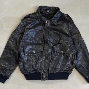 Può includere: Giacca bomber in pelle nera con collo, due tasche frontali e spalline. La giacca ha un orlo e polsini a coste. Un capo di abbigliamento esterno classico.
