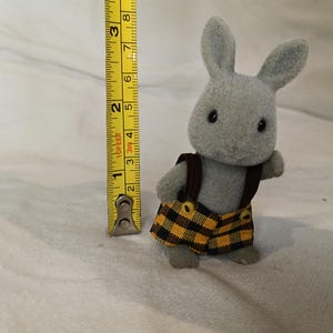 Puede incluir: Un conejo de juguete gris con un chaleco marr&oacute;n y pantalones cortos a cuadros amarillos y negros. El conejo mide aproximadamente 7,6 cm de altura, medido con una cinta m&eacute;trica amarilla.