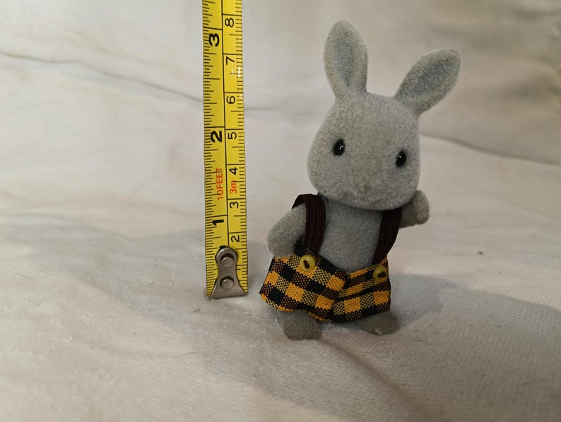 Puede incluir: Un conejo de juguete gris con un chaleco marr&oacute;n y pantalones cortos a cuadros amarillos y negros. El conejo mide aproximadamente 7,6 cm de altura, medido con una cinta m&eacute;trica amarilla.
