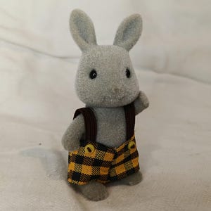 Puede incluir: Figura de conejo gris con un peto a cuadros amarillos y negros con correas marrones. El conejo tiene ojos negros y orejas erguidas. El juguete está sobre una superficie blanca.