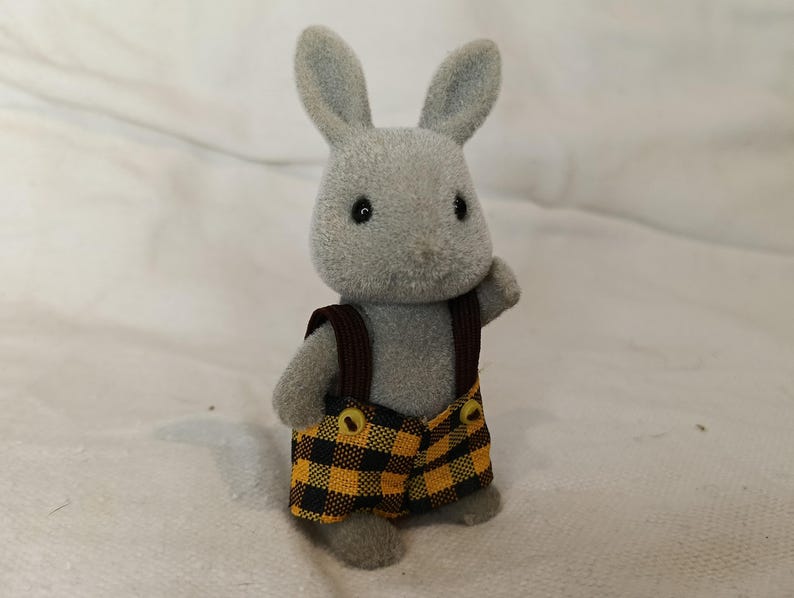 Puede incluir: Figura de conejo gris con un peto a cuadros amarillos y negros con correas marrones. El conejo tiene ojos negros y orejas erguidas. El juguete est&aacute; sobre una superficie blanca.