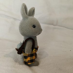 Puede incluir: Un conejo de juguete gris con ojos negros y una peque&ntilde;a cola esponjosa. El conejo lleva una bandolera marr&oacute;n y un pantal&oacute;n corto con un estampado de cuadros negros y amarillos. El juguete est&aacute; sobre una superficie blanca.