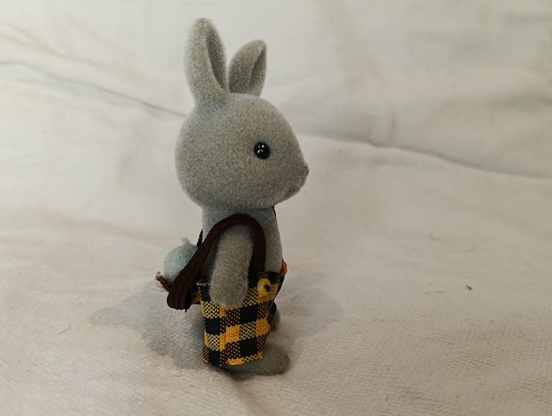 Puede incluir: Un conejo de juguete gris con ojos negros y una peque&ntilde;a cola esponjosa. El conejo lleva una bandolera marr&oacute;n y un pantal&oacute;n corto con un estampado de cuadros negros y amarillos. El juguete est&aacute; sobre una superficie blanca.
