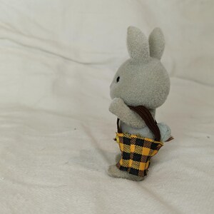 Puede incluir: Figura de conejo gris con una bolsa de correa marr&oacute;n con un patr&oacute;n de cuadros amarillos y negros. El conejo est&aacute; de pie sobre sus patas traseras, con las orejas levantadas. La bolsa cuelga de sus hombros.