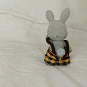 Puede incluir: Figura de conejo gris con un peto a cuadros amarillos y negros con tirantes marrones. El conejo tiene orejas largas y una peque&ntilde;a cola azul claro. El juguete est&aacute; sobre un fondo blanco.