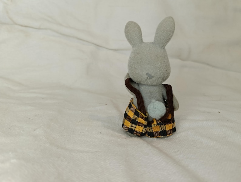 Puede incluir: Figura de conejo gris con un peto a cuadros amarillos y negros con tirantes marrones. El conejo tiene orejas largas y una peque&ntilde;a cola azul claro. El juguete est&aacute; sobre un fondo blanco.