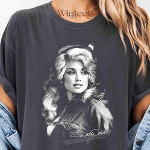 May include: A charcoal gray short-sleeve t-shirt featuring a black and white graphic print of a woman with voluminous hair. The graphic includes a signature at the bottom. The shirt has a crew neckline and a relaxed fit with rolled sleeves.