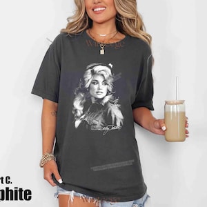 May include: A charcoal gray short-sleeve t-shirt featuring a black and white graphic print of Dolly Parton on the front. The graphic includes a portrait of the artist with her signature below it. The shirt has a crew neckline and a relaxed fit.