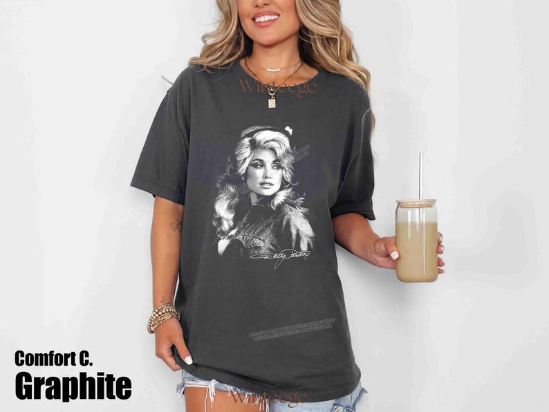 May include: A charcoal gray short-sleeve t-shirt featuring a black and white graphic print of Dolly Parton on the front. The graphic includes a portrait of the artist with her signature below it. The shirt has a crew neckline and a relaxed fit.