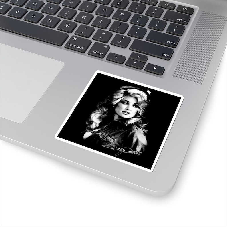May include: A square sticker featuring a black and white portrait of a person with voluminous blonde hair, set against a solid black background. The image includes a white signature at the bottom, and the sticker is placed on a laptop keyboard.