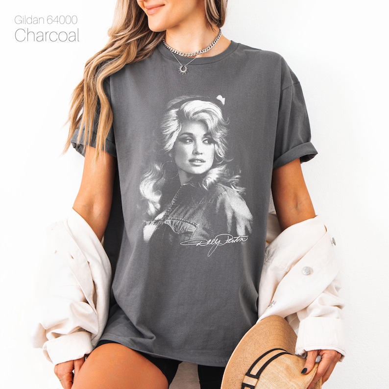 May include: A charcoal gray short-sleeve t-shirt featuring a white graphic print of Dolly Parton with her signature below the image. The text Gildan 64000 Charcoal is printed in the upper left corner of the image above the t-shirt.