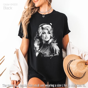 May include: A black Gildan 64000 t-shirt featuring a white graphic print of Dolly Parton with her signature below. The shirt has short sleeves and a crew neckline. The text Gildan 64000 Black is printed in the upper left corner of the image.