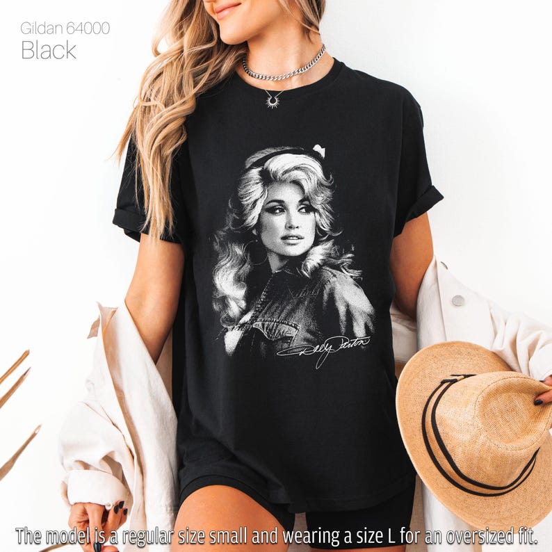 May include: A black Gildan 64000 t-shirt featuring a white graphic print of Dolly Parton with her signature below. The shirt has short sleeves and a crew neckline. The text Gildan 64000 Black is printed in the upper left corner of the image.