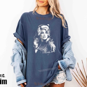 May include: A denim blue short-sleeve t-shirt featuring a white graphic print of a woman with voluminous hair. The design includes a signature at the bottom of the graphic. The shirt has a crew neckline and a relaxed fit, layered under a light blue denim jacket.