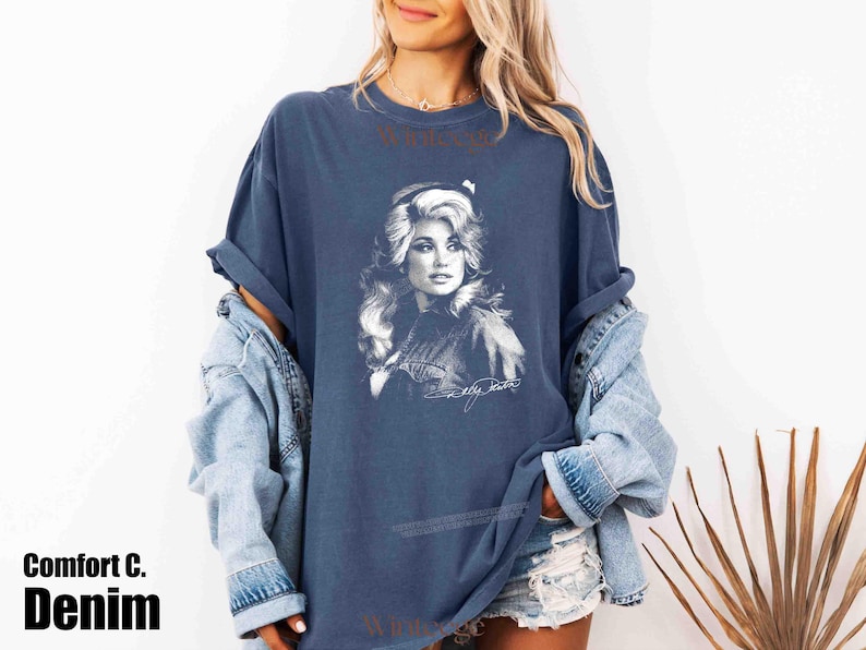 May include: A denim blue short-sleeve t-shirt featuring a white graphic print of a woman with voluminous hair. The design includes a signature at the bottom of the graphic. The shirt has a crew neckline and a relaxed fit, layered under a light blue denim jacket.