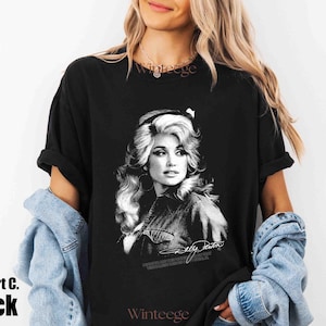 May include: A black short-sleeved t-shirt featuring a white graphic print of a portrait of Dolly Parton with her signature below the image. The shirt has a crew neckline and a relaxed fit, with the text Comfort C. Black visible in the corner.
