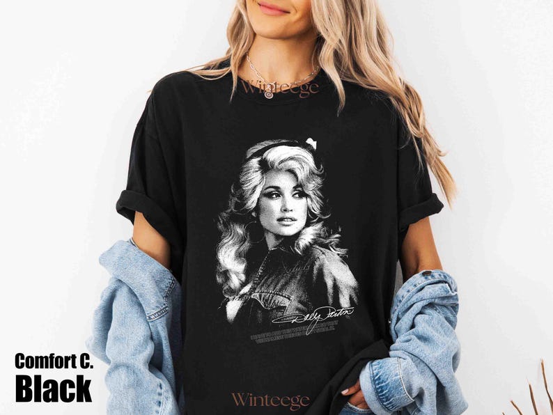 May include: A black short-sleeved t-shirt featuring a white graphic print of a portrait of Dolly Parton with her signature below the image. The shirt has a crew neckline and a relaxed fit, with the text Comfort C. Black visible in the corner.