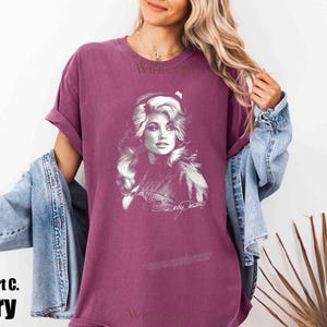 May include: A berry-colored short-sleeve t-shirt featuring a white graphic print of Dolly Parton on the front. The shirt has a crew neckline and a relaxed fit. The graphic includes a portrait of the artist and a signature below the image.
