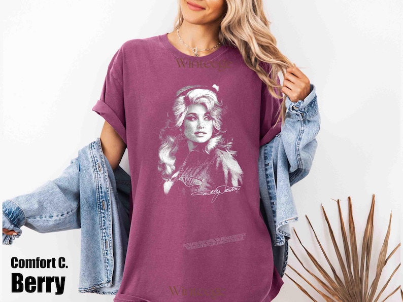 May include: A berry-colored short-sleeve t-shirt featuring a white graphic print of Dolly Parton on the front. The shirt has a crew neckline and a relaxed fit. The graphic includes a portrait of the artist and a signature below the image.