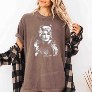 May include: A brown short-sleeved t-shirt featuring a white graphic print of a woman with long wavy hair. The shirt includes the text "Dolly Parton" in a cursive script below the portrait. A black and tan plaid long-sleeved shirt is worn over it.