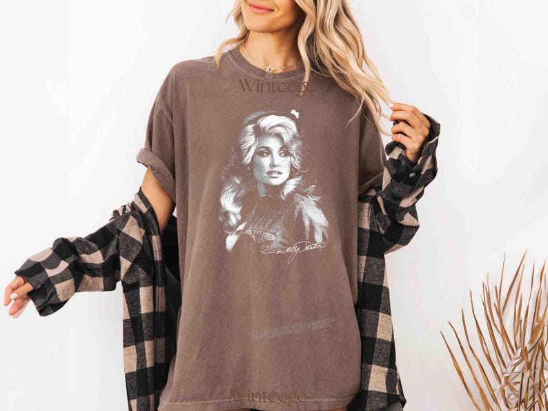 May include: A brown short-sleeved t-shirt featuring a white graphic print of a woman with long wavy hair. The shirt includes the text "Dolly Parton" in a cursive script below the portrait. A black and tan plaid long-sleeved shirt is worn over it.