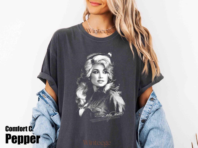 May include: A charcoal gray short-sleeve t-shirt featuring a black and white graphic print of a woman with voluminous hair. The graphic includes a signature at the bottom. The shirt has a crew neckline and a relaxed fit with rolled sleeves.