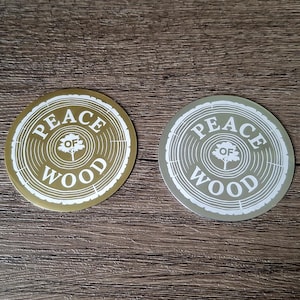 Könnte beinhalten: Zwei runde Aufkleber mit einer weißen Baumringgrafik und dem Text "PEACE OF WOOD" sowie einem kleinen Baumsymbol in der Mitte. Ein Aufkleber hat einen metallisch goldenen Hintergrund, der andere einen metallisch silbernen, beide mit weißer Tinte.