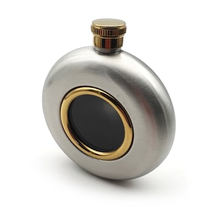 Könnte beinhalten: Eine Edelstahl-Taschenflasche mit gebürsteter Oberfläche. Sie hat einen goldfarbenen Rand um eine schwarze Mitte und einen goldfarbenen Deckel. Die Flasche ist rund und hat ein modernes Design.