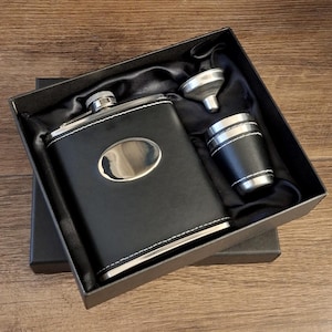 May include: A black leather-covered flask with a silver lid and a silver oval plate on the front. The flask is in a black box with a black velvet lining. The box also contains a silver funnel and a silver shot glass with a black leather cover.