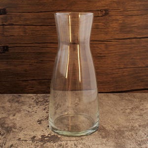 Op de afbeelding: Een heldere glazen karaf met een brede basis, zacht gebogen zijkanten en een uitlopende bovenkant. De karaf is leeg en staat op een getextureerd oppervlak, met een houten achtergrond. Het ontwerp is eenvoudig en elegant, geschikt voor het serveren van dranken.