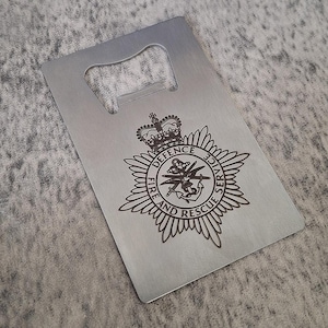 May include: A rectangular, brushed stainless steel credit card-sized bottle opener. The surface features a laser-engraved emblem of the Defence Fire and Rescue Service, which includes a central anchor and propeller design topped by a royal crown.