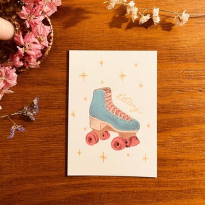 Puede incluir: Un patín de ruedas azul con ruedas rosas y cordones blancos. El patín está sobre un fondo blanco con estrellas doradas y la palabra "rolling" escrita en cursiva negra.